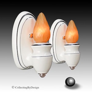 May include: A pair of wall sconces featuring white ceramic oval backplates with thin black trim and matching white ceramic bases. Each sconce is topped with a frosted orange glass shade shaped like a flickering flame, emitting a warm glow.