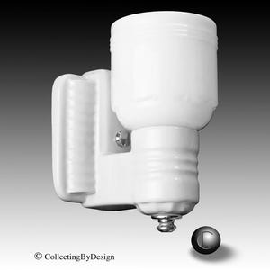 May include: A white ceramic wall-mounted light fixture featuring a cylindrical shade and a ribbed base. The fixture includes a small metallic screw component at the bottom and a circular logo with the letter C and the text CollectingByDesign.