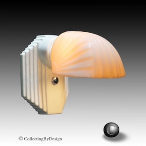 May include: A wall-mounted light fixture featuring a white, vertically ribbed, stepped-back base and a semi-circular, frosted glass shade shaped like a seashell. The shade emits a warm, soft glow, and a small metal screw is visible on the base.