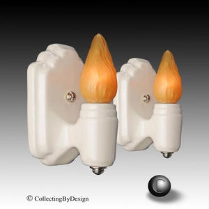 May include: A pair of wall sconces featuring white ceramic backplates with a stepped, geometric design. Each sconce holds a single flame-shaped, amber-colored glass shade with a textured, swirling surface. A small metal screw is visible on each.