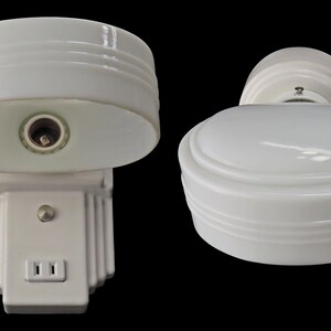 Puede incluir: Dos apliques de pared blancos de estilo vintage con un dise&ntilde;o aerodin&aacute;mico. Los apliques presentan una pantalla redondeada con estr&iacute;as horizontales y una base rectangular con una toma de corriente. Los accesorios est&aacute;n sobre un fondo negro.