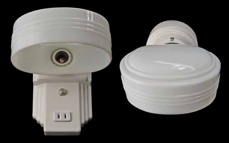 Puede incluir: Dos apliques de pared blancos de estilo vintage con un dise&ntilde;o aerodin&aacute;mico. Los apliques presentan una pantalla redondeada con estr&iacute;as horizontales y una base rectangular con una toma de corriente. Los accesorios est&aacute;n sobre un fondo negro.