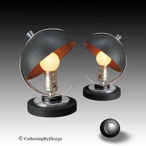 May include: Two matching black and silver table lamps with a unique design. Each lamp features a black, semi-circular shade with a copper-colored interior, a silver frame, and a round base. The lamps have a vintage, mid-century modern aesthetic.