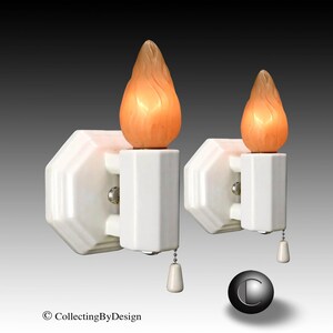 May include: Two white wall sconces with flame-shaped light bulbs. Each sconce has a pull chain and a decorative, octagonal base. The bulbs are orange and yellow, resembling a flickering flame. The background is a gradient of gray.