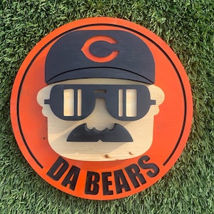May include: A round wooden sign with an orange background and a black outline. The sign features a cartoon illustration of a person wearing a black hat with a red "C" on it, sunglasses, and a mustache. The text "DA BEARS" is written in black letters on the bottom of the sign.