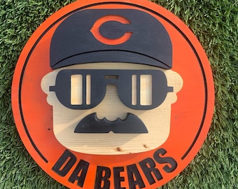May include: A round wooden sign with an orange background and a black outline. The sign features a cartoon illustration of a person wearing a black hat with a red "C" on it, sunglasses, and a mustache. The text "DA BEARS" is written in black letters on the bottom of the sign.