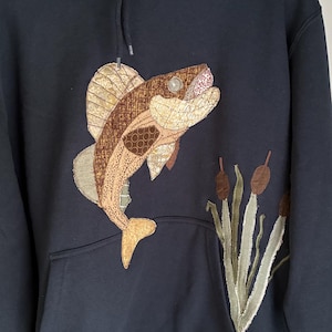 May include: A dark charcoal gray hooded sweatshirt featuring a large, multi-textured fabric appliqué of a fish leaping upward. To the right of the fish, there is a fabric appliqué of green cattail plants with brown heads. The hoodie has a front pocket.