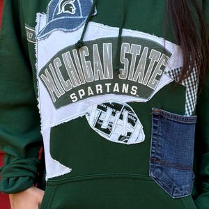 May include: A dark green hooded sweatshirt featuring a large Michigan State Spartans pennant appliqué, a denim baseball cap patch, a football-shaped fabric piece, and a dark blue denim pocket sewn onto the front with frayed white fabric edges.