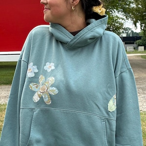 May include: A dusty blue hooded sweatshirt featuring a large front kangaroo pocket and decorative fabric flower appliqués on the chest. The floral patches display intricate paisley and swirling patterns in shades of blue, yellow, and white.