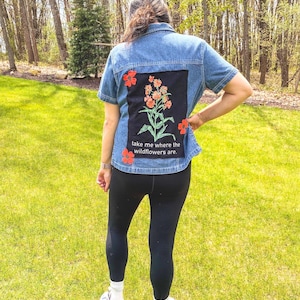 May include: A denim jacket with short sleeves, featuring a black patch on the back with a floral design in orange and green. The text "take me where the wildflowers are." is printed below the design. Red flower appliqués are placed around the patch.