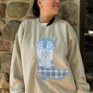 May include: A beige crewneck sweatshirt featuring a large, central appliqué in the shape of a cowboy boot. The boot design is split into two sections: a white upper portion with a blue snowflake pattern and a lower portion with a blue plaid pattern.
