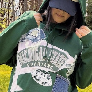 May include: A dark green hooded sweatshirt featuring a large Michigan State Spartans applique on the front. The design includes frayed fabric patches, a denim pocket, and a football graphic. The hoodie has matching green drawstrings.
