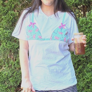 May include: A white short-sleeve t-shirt featuring a printed turquoise bikini top graphic with pink flamingo patterns and small pink bows on the straps, paired with navy blue and white horizontal striped shorts, a straw hat, and black sunglasses.