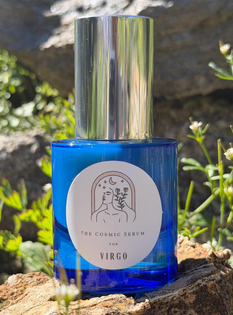 Puede incluir: Una botella de vidrio azul con tapa plateada. La botella tiene una etiqueta blanca con una ilustraci&oacute;n y el texto "THE COSMIC SERUM for VIRGO". La botella est&aacute; sobre un fondo natural de rocas y vegetaci&oacute;n.