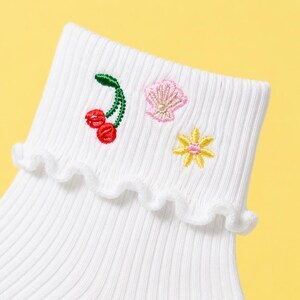 May include: White ribbed socks with a frilled cuff, featuring embroidered cherries, a seashell, and a flower. The cherries are red, the seashell is pink, and the flower is yellow. The socks are set against a yellow backdrop.