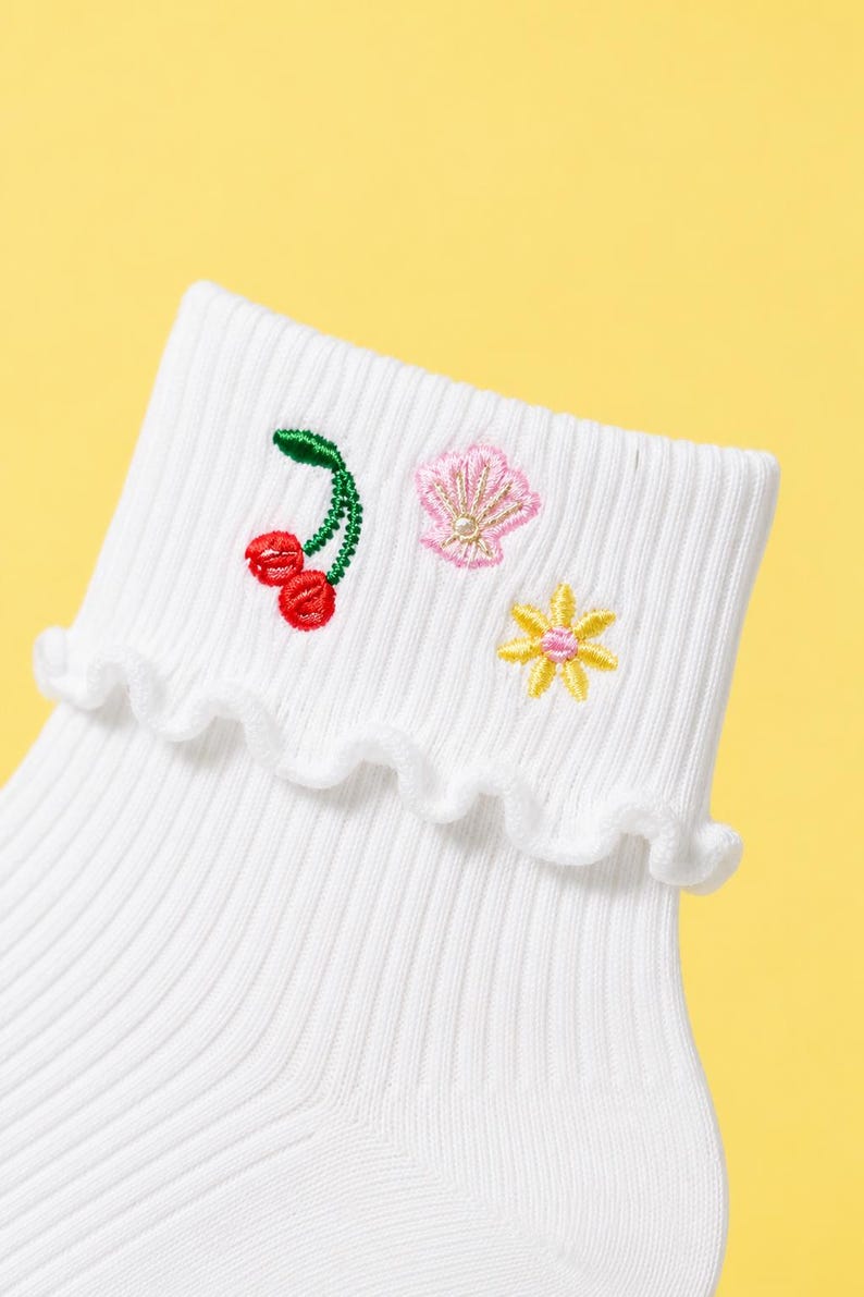 May include: White ribbed socks with a frilled cuff, featuring embroidered cherries, a seashell, and a flower. The cherries are red, the seashell is pink, and the flower is yellow. The socks are set against a yellow backdrop.