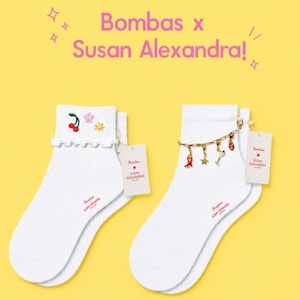 May include: Two pairs of white ankle socks with the text "Bombas x Susan Alexandra!" in pink. One pair has cherry and flower embroidery, the other has a charm bracelet with red and gold charms. Each sock has a tag.