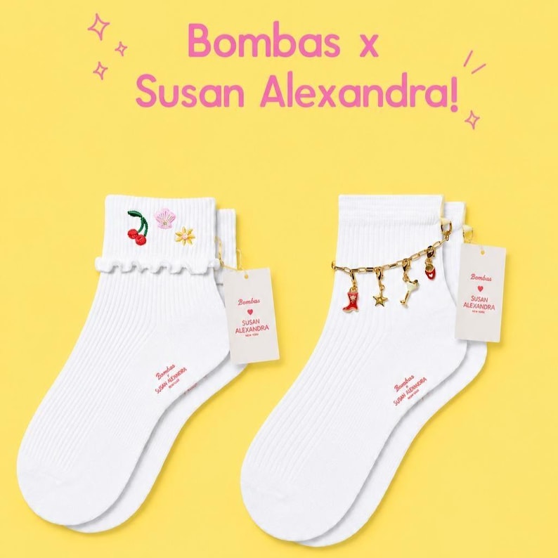 May include: Two pairs of white ankle socks with the text "Bombas x Susan Alexandra!" in pink. One pair has cherry and flower embroidery, the other has a charm bracelet with red and gold charms. Each sock has a tag.