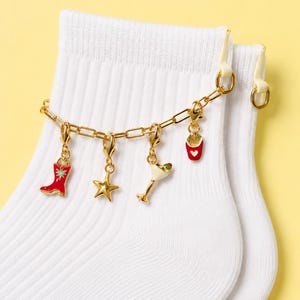 May include: White socks with a gold chain charm bracelet. The bracelet features charms including a red cowboy boot, a gold star, a martini glass, and a container of fries. The background is a solid yellow.