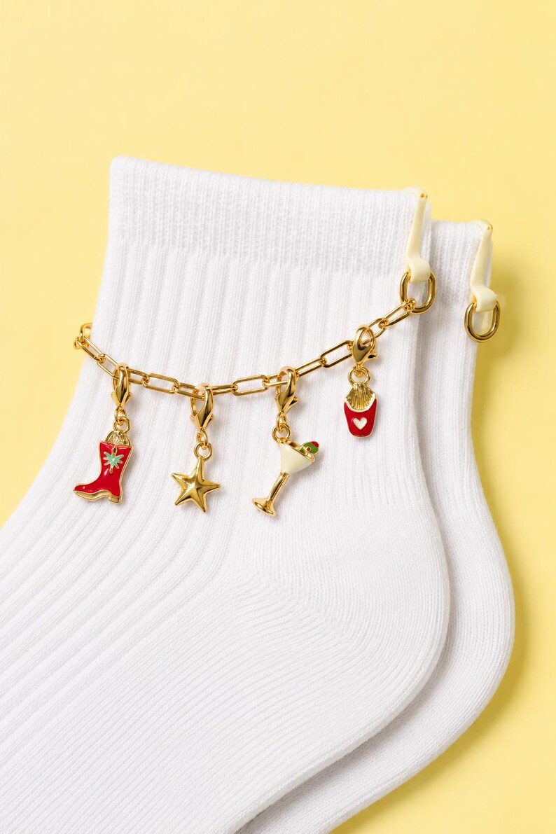 May include: White socks with a gold chain charm bracelet. The bracelet features charms including a red cowboy boot, a gold star, a martini glass, and a container of fries. The background is a solid yellow.