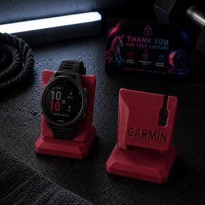 May include: A black Garmin smartwatch with a black band rests on a red stand. The stand is next to another red stand with the Garmin logo. A thank you card with a unicorn graphic is also visible.