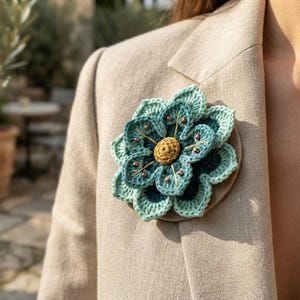 May include: A crochet flower brooch featuring layered petals in shades of light and dark teal, accented with a central yellow textured knot and small metallic beads. The floral piece is attached to a circular base and pinned to a beige blazer.