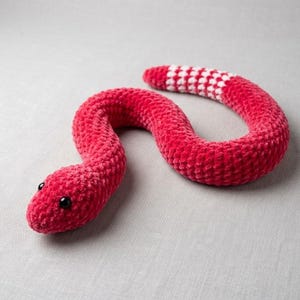 Può includere: Un serpente di peluche realizzato in filato rosso vivace con una finitura morbida e testurizzata. Il serpente presenta due piccoli occhi in plastica nera e una sezione della coda distinta con un motivo a scacchi alternato bianco e rosso.