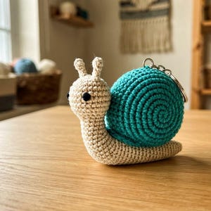 Puede incluir: Un llavero de caracol tejido a ganchillo con un cuerpo de color crema, dos pequeñas antenas y ojos de cuentas negras. El caracol tiene una concha redonda en espiral de color azul verdoso vibrante, con una anilla de metal plateado en la parte superior.