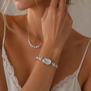 May include: A silver watch with an oval white face and a floral design on the strap. A matching floral necklace is also visible. The woman is wearing a white camisole with lace trim. The jewellery is delicate and elegant.