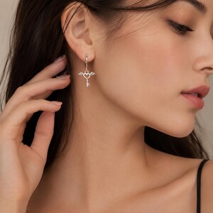 Puede incluir: Pendientes colgantes plateados. Cada pendiente presenta un coraz&oacute;n con alas y un dije de llave. Los pendientes est&aacute;n sujetos a un peque&ntilde;o aro que atraviesa la oreja. El dise&ntilde;o es delicado y detallado.