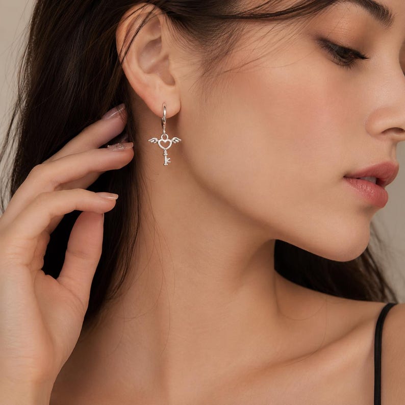 Puede incluir: Pendientes colgantes plateados. Cada pendiente presenta un coraz&oacute;n con alas y un dije de llave. Los pendientes est&aacute;n sujetos a un peque&ntilde;o aro que atraviesa la oreja. El dise&ntilde;o es delicado y detallado.