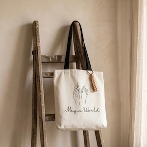 Peut inclure: Sac fourre-tout crème avec des anses noires et un pompon en cuir marron. Le sac présente un dessin au trait noir de mains tenant une plante et les mots "Magic World". Le sac est suspendu à une échelle en bois.