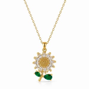 May include: A gold-toned necklace featuring a sunflower pendant with clear marquise-cut stones as petals, a textured gold center, and a gold stem with two teardrop-shaped green stones as leaves, suspended from a delicate gold-toned chain.