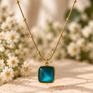 May include: A gold-toned necklace featuring a delicate chain with evenly spaced small beads and a square pendant. The pendant contains a smooth, translucent teal-colored stone with a distinct cat's eye effect centered in a gold-colored frame.