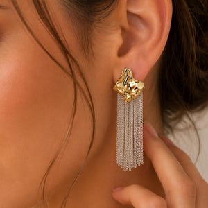 May include: Gold-tone earrings with a textured, abstract design and dangling silver-tone chain fringe. The earrings are worn on the ear, showcasing the intricate details and contrasting textures of the jewelry.