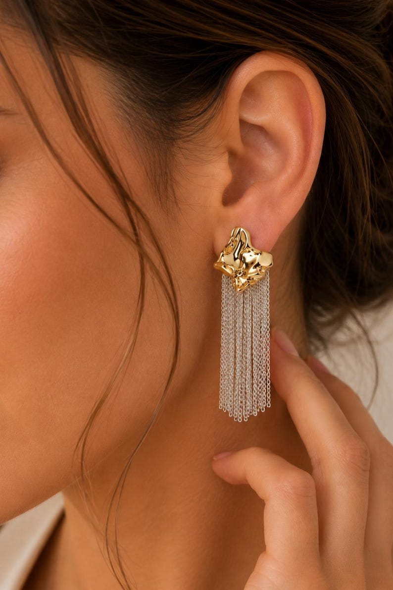 May include: Gold-tone earrings with a textured, abstract design and dangling silver-tone chain fringe. The earrings are worn on the ear, showcasing the intricate details and contrasting textures of the jewelry.