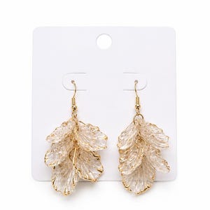 May include: A pair of dangle earrings with a leaf-like design. The earrings feature multiple layers of clear, textured, petal-shaped elements with gold-colored edges and hooks. They are displayed on a white card.
