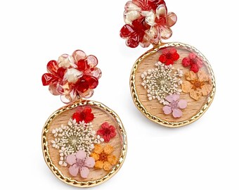 May include: A pair of round earrings featuring dried flowers encased in clear resin. The earrings have a gold-colored border and a cluster of red and clear beads at the top. The flowers are in shades of red, white, pink, and orange.