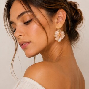 May include: Close-up of a woman wearing gold-colored earrings. The earrings are in the shape of a flower with a cluster of small, light pink beads. The woman is wearing a white off-the-shoulder top.