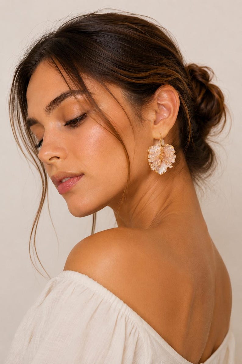 May include: Close-up of a woman wearing gold-colored earrings. The earrings are in the shape of a flower with a cluster of small, light pink beads. The woman is wearing a white off-the-shoulder top.