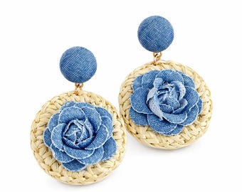May include: A pair of dangle earrings featuring a circular woven straw base topped with a blue denim fabric flower. Each earring has a round blue denim stud post at the top, connected to the straw disc by a small gold-toned metal link.