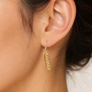 May include: Gold-colored wheat-shaped earrings. The earrings are hanging from a person's ear. The earrings have a hook design and a detailed wheat stalk. The earrings are made of a shiny, metallic material.