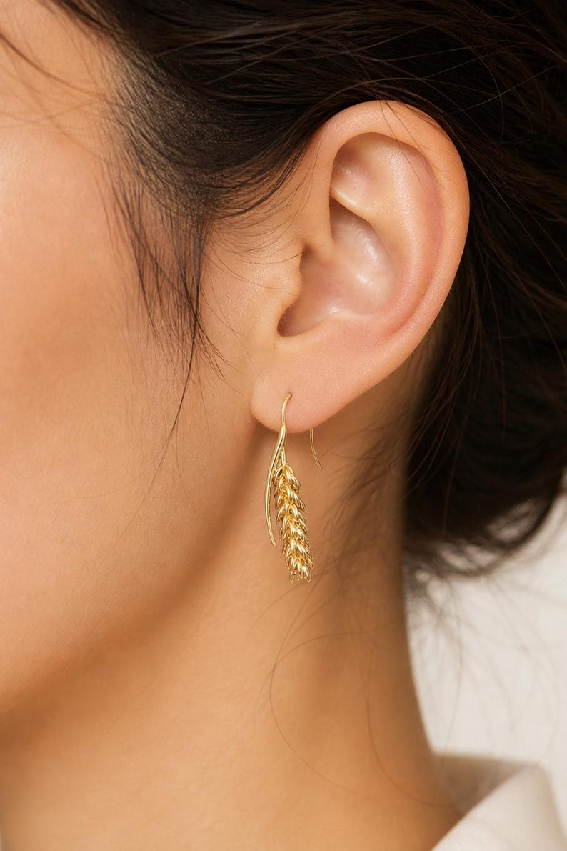 May include: Gold-colored wheat-shaped earrings. The earrings are hanging from a person's ear. The earrings have a hook design and a detailed wheat stalk. The earrings are made of a shiny, metallic material.