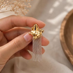 May include: A gold-colored earring with a textured, organic shape and a cascade of silver-colored chain fringe. The earring is held up against a neutral background, showcasing its design and details. This piece is a statement accessory.