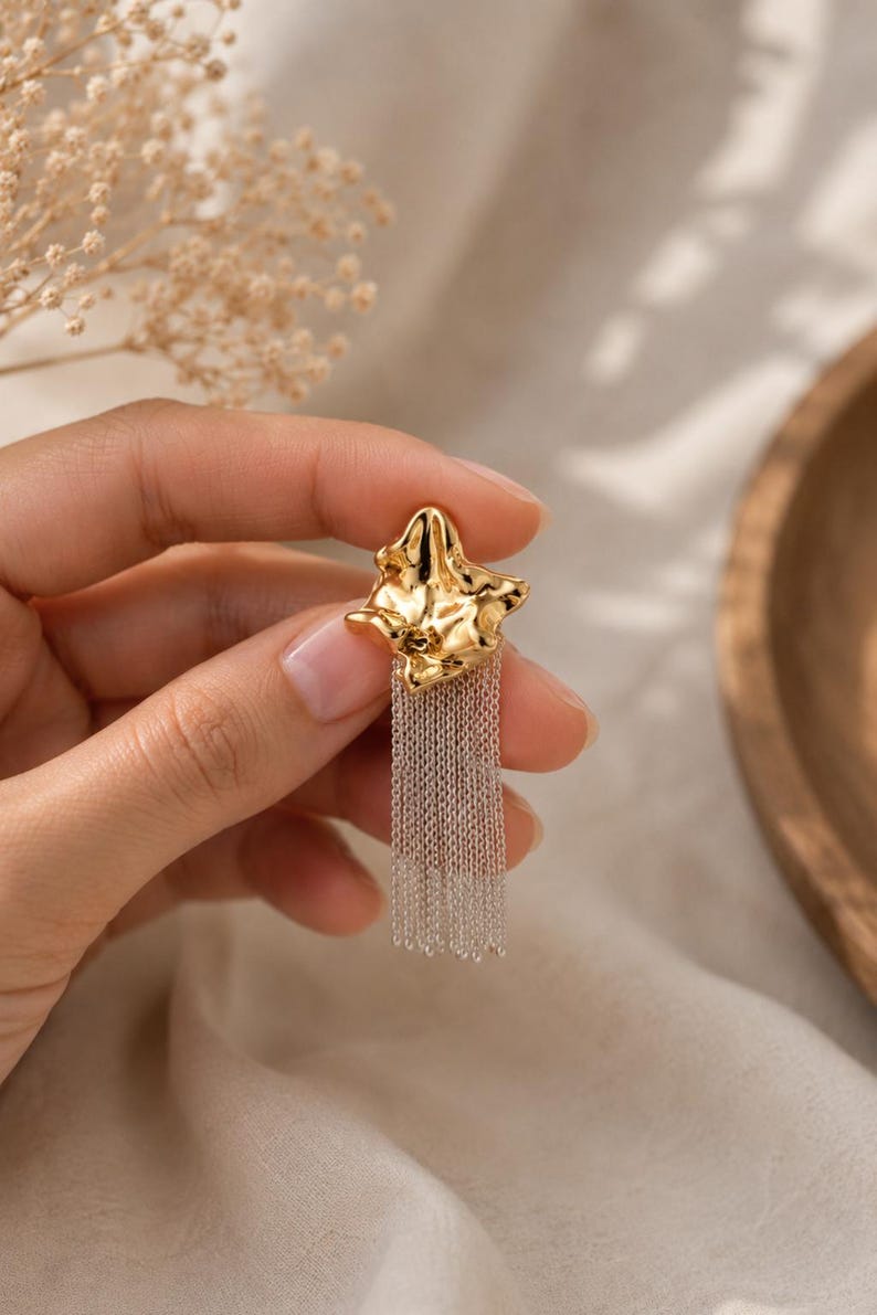 May include: A gold-colored earring with a textured, organic shape and a cascade of silver-colored chain fringe. The earring is held up against a neutral background, showcasing its design and details. This piece is a statement accessory.