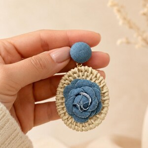May include: A dangle earring featuring a circular woven rattan frame with a blue denim fabric flower at the center. The earring has a matching blue denim fabric-covered stud top connected by a small gold-tone metal jump ring.