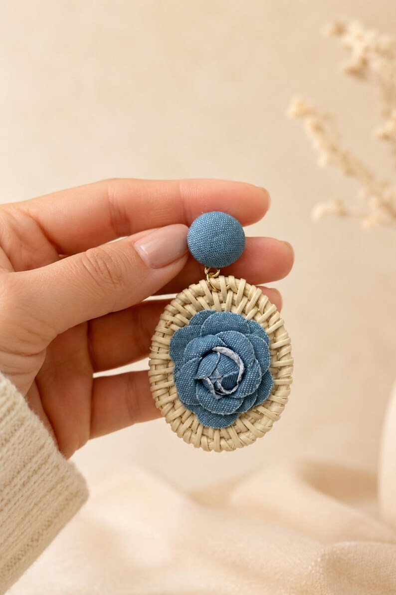 May include: A dangle earring featuring a circular woven rattan frame with a blue denim fabric flower at the center. The earring has a matching blue denim fabric-covered stud top connected by a small gold-tone metal jump ring.