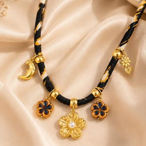 May include: A necklace featuring a black fabric cord with a yellow and white pattern, adorned with gold-toned charms including a crescent moon, a small flower, two dark blue enamel flowers, and a central gold flower with a white pearl center.