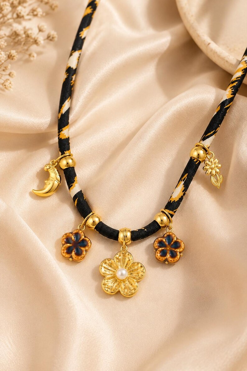 May include: A necklace featuring a black fabric cord with a yellow and white pattern, adorned with gold-toned charms including a crescent moon, a small flower, two dark blue enamel flowers, and a central gold flower with a white pearl center.