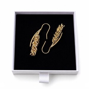 May include: A pair of gold-colored earrings shaped like wheat stalks, presented in a white box with a black interior. The earrings have a hook design for pierced ears and are displayed against a contrasting black background.